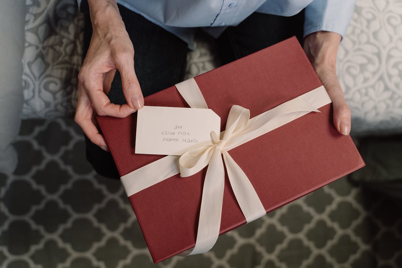 Offerings Close-up of hands holding a red gift box with a white ribbon and a message tag.